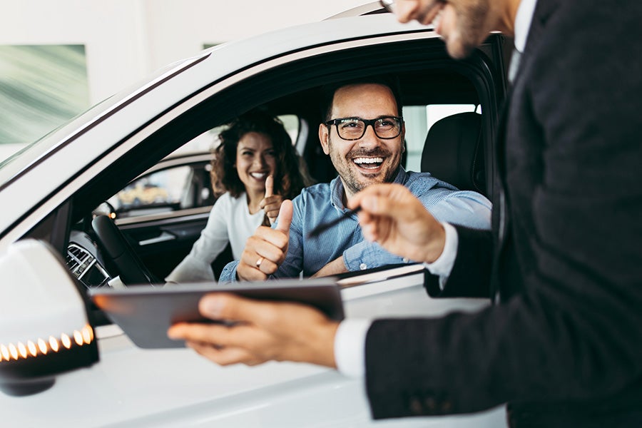 Two happy people in a car speaking to a salesman at a dealership
